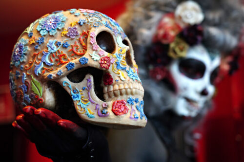 A black painted hand covered in red paint holds a skull figure decorated with floral patterns in shades of red, orange, yellow, green, blue, and purple. Painted white with black accents to resemble a skull, the face—and also gray hair and flower headband—of the person holding the skull are blurry in the background.Cindy Ord/Getty Images