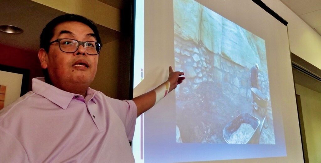 A man in a purple short-sleeved shirt and glasses points to a PowerPoint slide of an archaeological excavation featuring stone walls, two wheelbarrows, and a man wearing a hard hat.