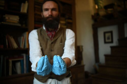 A bearded man in a vest and red scarf holds up his two hands in loose blue gloves, with a small tooth in each hand.