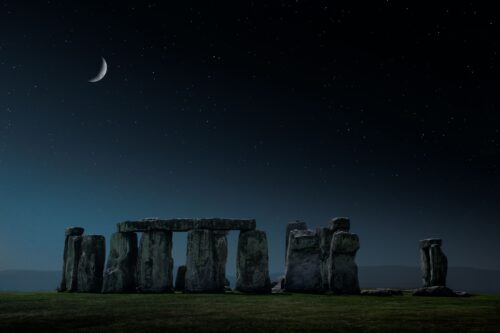 Massive vertical and horizontal stones stand in a field at night, with the sliver of a silver moon above.