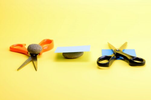 Two pairs of scissors, two rocks, and two slips of paper rest on a pale-yellow surface.