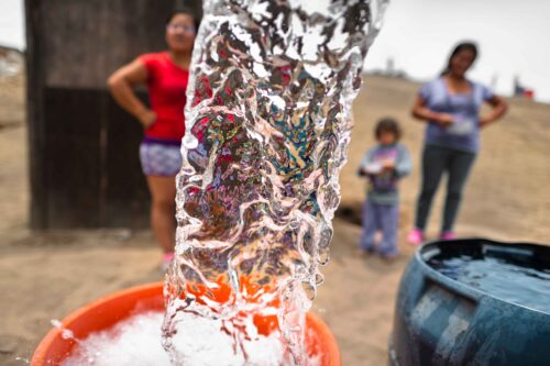 A stream of clear water pours into an orange bucket. Behind it, two slightly out-of-focus women and a child look on.