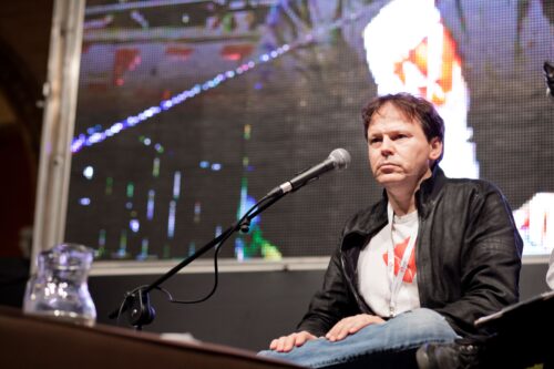 A man in a t-shirt, jacket, and jeans sits comfortably with a leg folded, on a stage. A microphone extends in front of him.