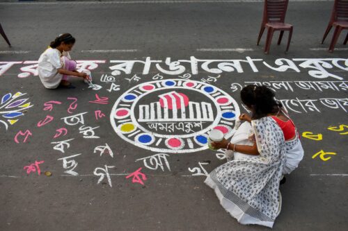 Two people crouch down to create colorful chalk art on a street. Intricate designs and language characters surround a central circular moti