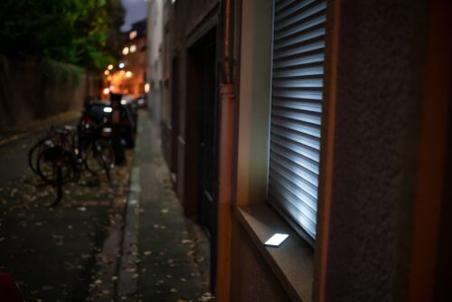 On a quiet street at night, a small, glowing rectangular device rests on the sill of a stall window shuttered with a corrugated metal cover.