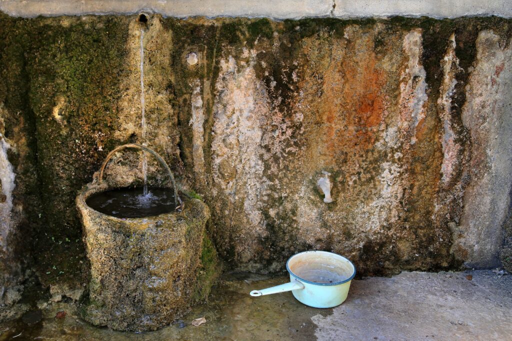 An old bucket coated in multicolored limestone sits in front of a limestone-covered wall out of which protrudes a faucet dripping water that falls into the bucket.