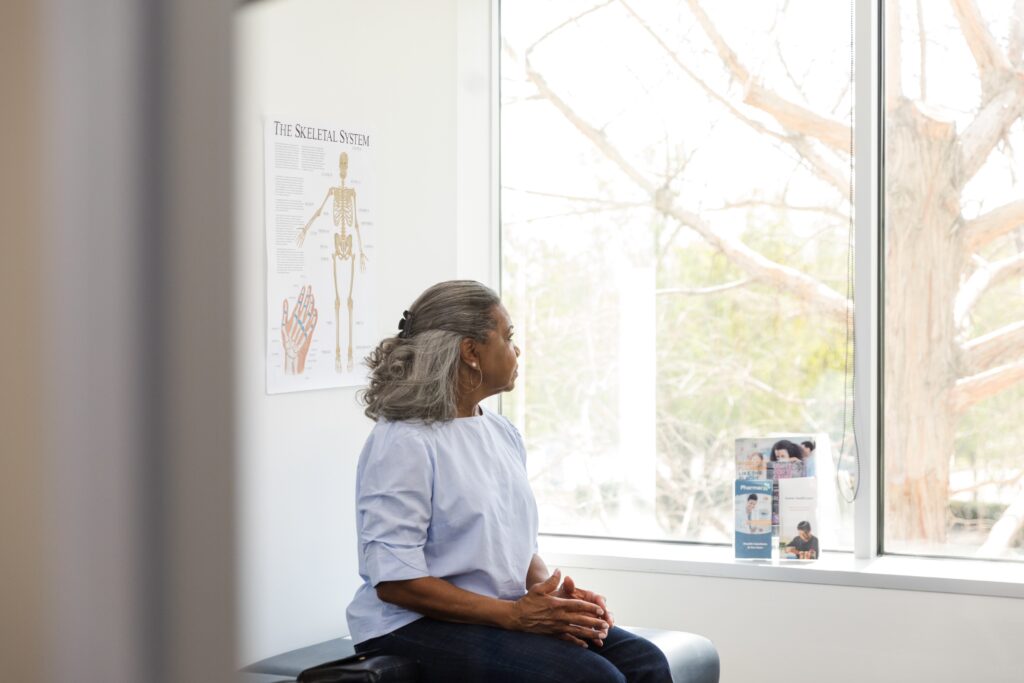 A gray-haired woman sits on an exam table in a light-filled doctor’s office and looks out a window.