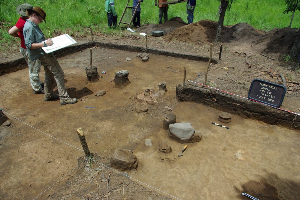 Two people stand inside a rectangular dirt plot that has been dug below ground. One of them takes notes on a clipboard. The brown, earthy site is surrounded by piles of dirt and green grass.