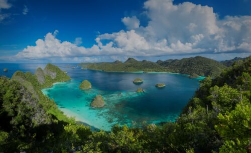 A U-shaped cove encircled by rugged peaks covered in green forest opens to an ocean in varying shades of blues that extends to the horizon where colossal white clouds sit.