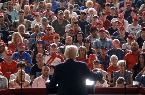 A photo shows the back of a person in a dark suitcoat standing at a podium and speaking to a large, captive, seated audience. Most onlookers wear red MAGA hats and Trump T-shirts.