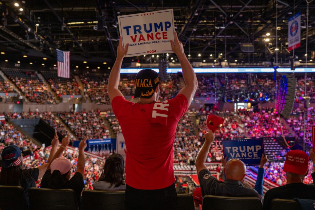 A man in a dark MAGA hat and red T-shirt holds up a sign that reads “Trump Vance Make America Great Again” in an arena filled with thousands of people also wearing MAGA gear. Many people are cheering and waving signs.