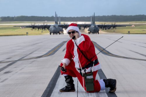 A person dressed in a red Santa Claus suit with white trim kneels on a military runway while speaking into a handheld radio. In the background are two rows of large military airplanes.