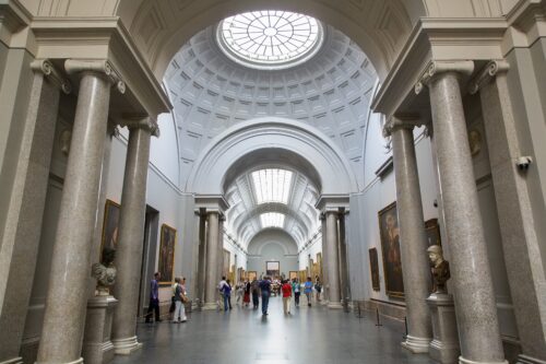 People walk through a long, wide museum hallway with high arched ceilings and skylights framed by tall tan columns.