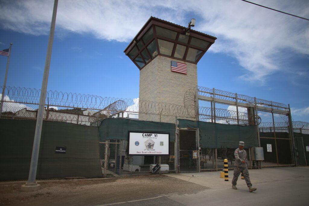 A person in army fatigues walks in front of a looming surveillance tower flanked by high fences topped with barbed wire.