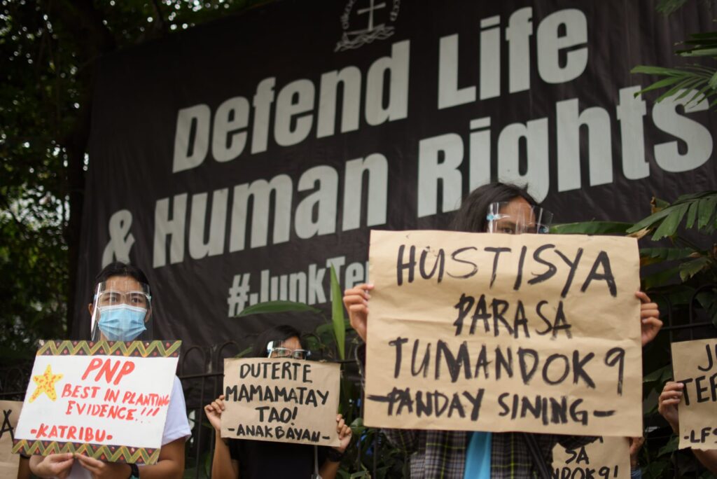 In front of a black sign with white letters that reads "Defend Life & Human Rights," people hold hand-made signs.