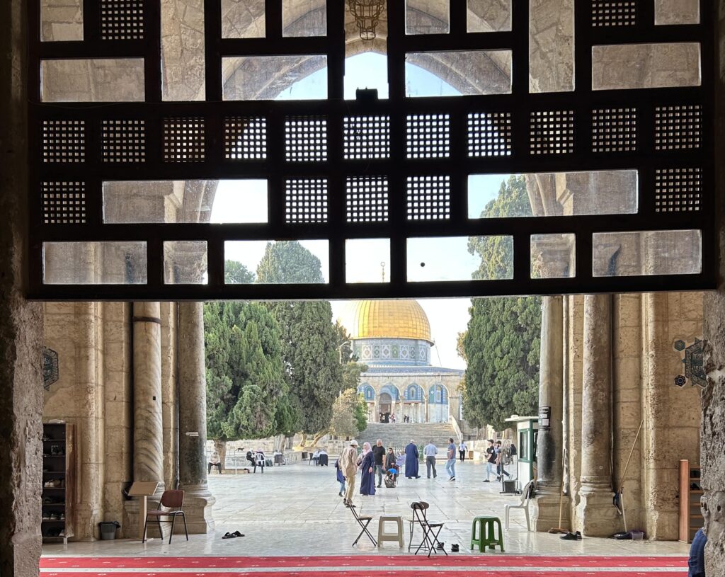 A golden dome rises in the background atop a mosque. The image was taken through a doorway that includes a dark screen framing the sidewalk and moque.