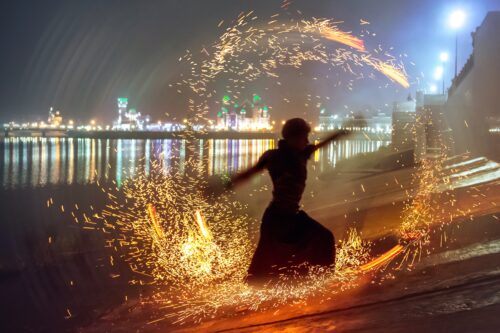 A person swings flame in a circle around them at night, with lights of a city in the background.