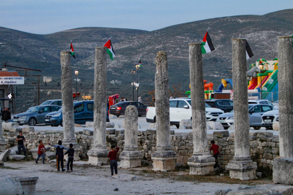 Children run in front of an ancient stone wall and columns adorned with green, red, white, and black flags. Cars are parked in an adjacent lot that looks out onto gently rolling green and brown hills.