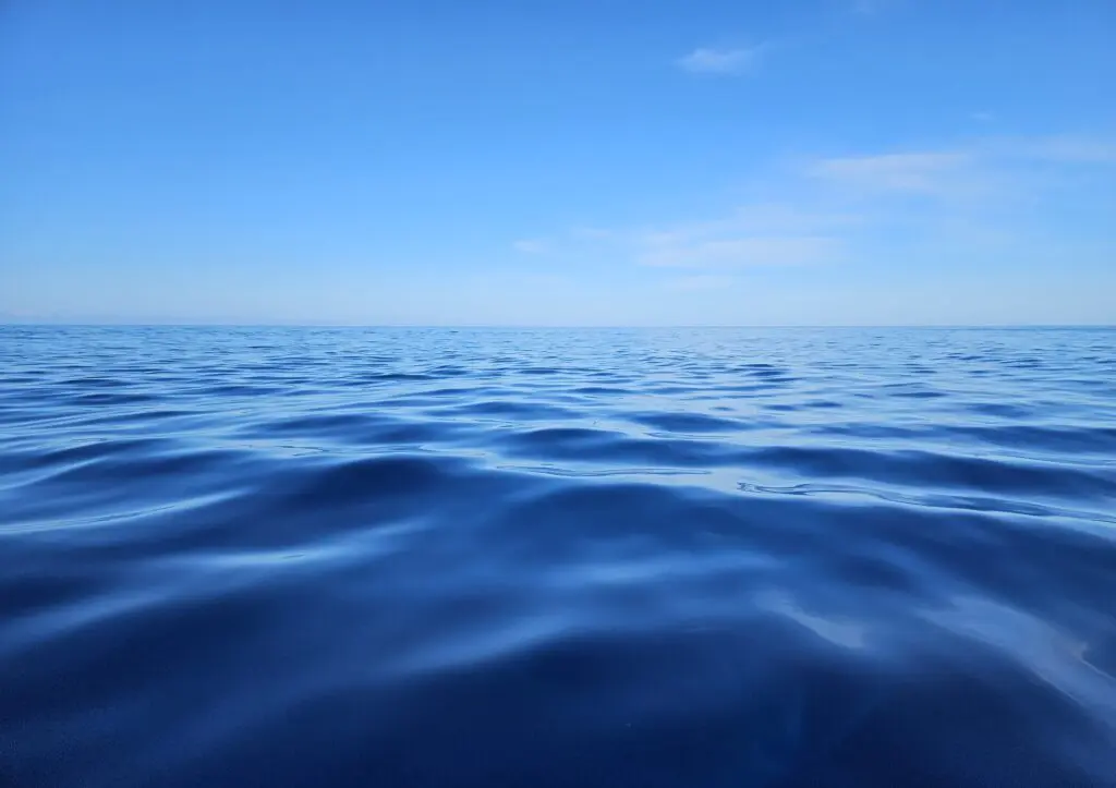 Beneath a blue sky with a few wispy white clouds, the glassy, rippling deep-blue surface of the ocean stretches to the horizon.